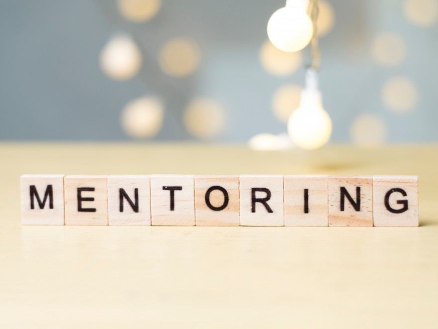 Photo has Scrabble letters that spell out the word mentoring with lights in the background.